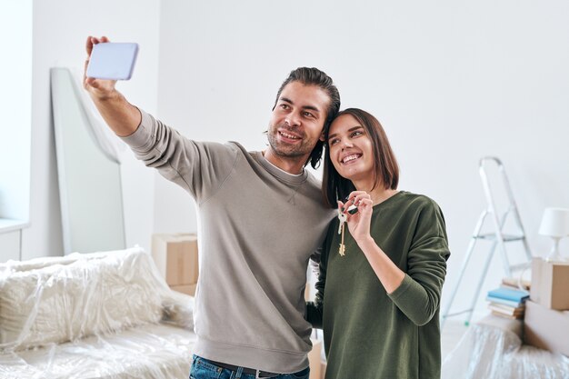 Jonge man met smartphone selfie maken met zijn gelukkige vrouw die de sleutel ingedrukt houdt naar hun nieuwe huis of flat terwijl ze in de woonkamer staan