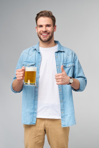 Jonge Man met spijkerbroek shirt bedrijf glas bier staande over grijze muur te houden.