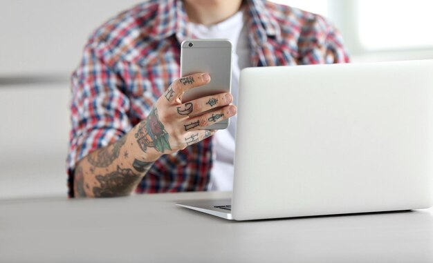 Jonge man met tatoeage met behulp van laptop en mobiele telefoon aan tafel
