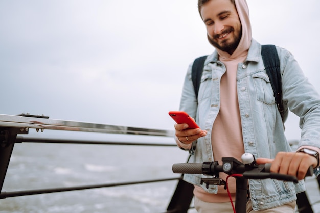 Jonge man met telefoon elektrische scooter rijden op een pier in de buurt van zee Ecologisch transportconcept