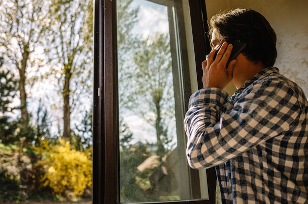 Jonge man met vrijetijdskleding die 's ochtends op een mobiele telefoon praat bij een raam met kopieerruimte