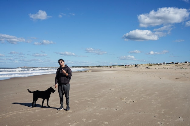 Jonge man met zwarte hond op het strand zwaaiend naar de camera. Het rock-'n-roll-symbool maken met zijn hand.