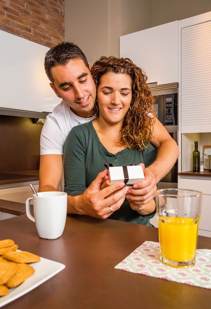 Jonge man omhelst en geeft een geschenkdoos aan zijn verraste vriendin tijdens het ontbijt in de keuken thuis