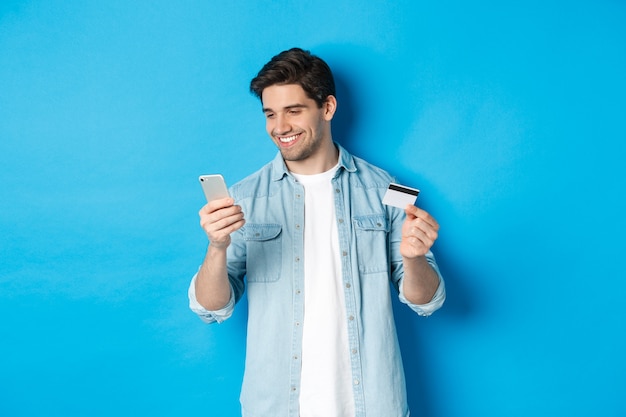 Jonge man online winkelen met mobiele applicatie, smartphone en creditcard vasthoudend, staande over blauwe achtergrond