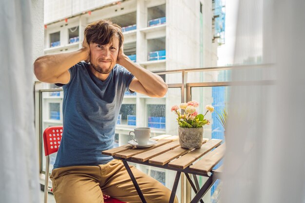 Jonge man op het balkon geïrriteerd door de bouwwerkzaamheden buiten Geluidsconcept Luchtvervuiling door bouwstof