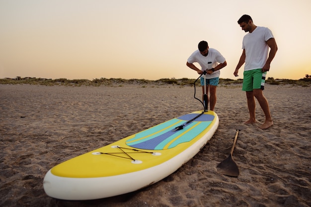 Jonge man opblazen paddle sup board op het strand bij zonsopgang