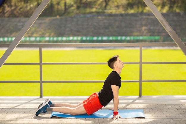 Jonge man opleiding yoga buitenshuis. Sportieve man maakt rekoefeningen op een blauwe yogamat, op het sportveld.