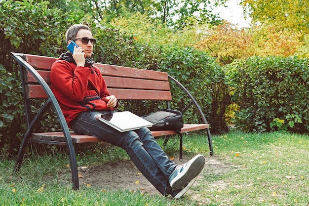 Jonge man praten op mobiele telefoon tijdens afstandswerk op laptopcomputer zittend in stadspark in dag Hipster man belt via mobiele telefoon en leest informatie van ultrabook oplossen van problemen