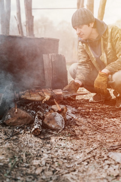 Jonge man survivalist kookt gebraden kippenvlees eten gebakken op de grill op smeulende kolen of sintels van een kampvuur op de grond barbecue in kampeeromstandigheden platteland en wild rustiek leven flare