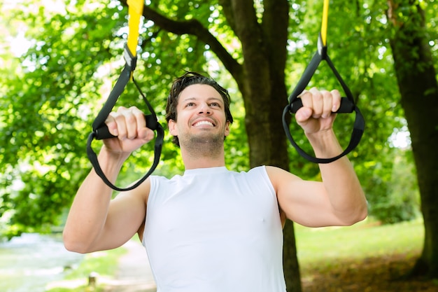 Jonge man trainen met schorsing trainer slinger in stadspark onder bomen van de zomer voor sport fitness