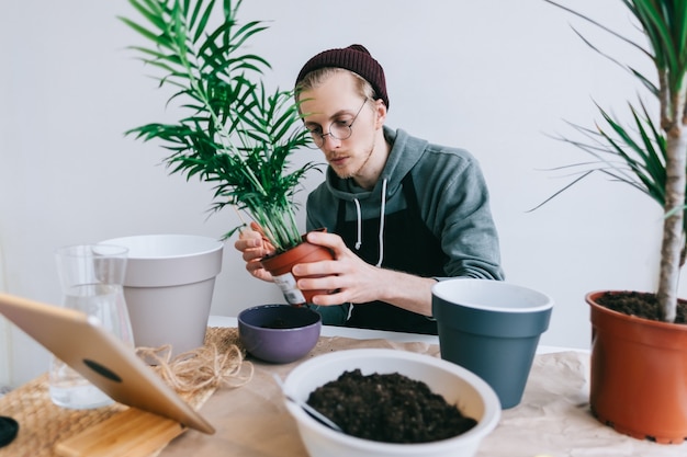 Jonge man tuinman in brillen plant in potten op de witte houten tafel overplanten en met behulp van tabletcomputer