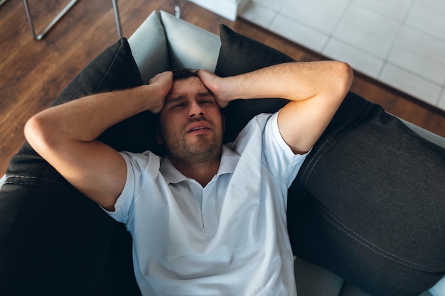 Jonge man tv kijken in zijn eigen appartement. Hoofdpijn, pijn of kater. Hand in hand op zijn hoofd en lijden.