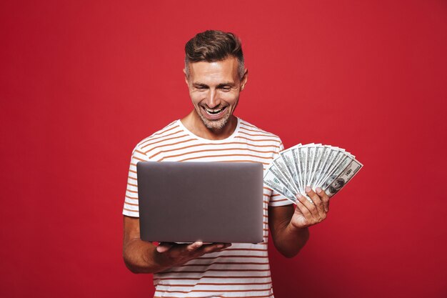 jonge man van 30 in gestreept t-shirt glimlachend terwijl hij een fan van geldbankbiljetten en een laptop vasthoudt die op rood is geïsoleerd