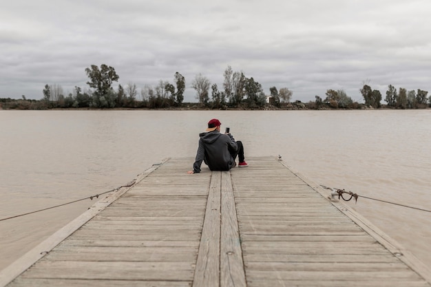 Jonge man van achteren het dragen van een grijze trui en een rode pet met behulp van een smartphone en het nemen van foto's zittend op een pier naast een rivier op een bewolkte dag