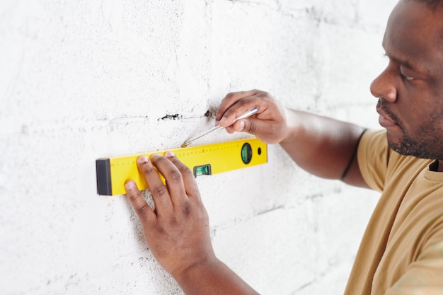 Jonge man van Afrikaanse etniciteit met potloodlijn op wit geschilderde bakstenen muur tijdens het gebruik van handgereedschap voor het meten van niveau tijdens woningrenovatie