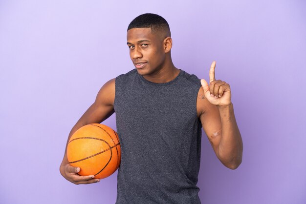 Jonge man van de basketbalspeler die op purpere muur wordt geïsoleerd die en een vinger in teken van het beste opheft