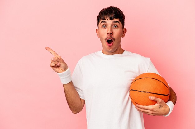 Jonge man van gemengd ras die basketbal speelt geïsoleerd op een roze achtergrond die naar de zijkant wijst