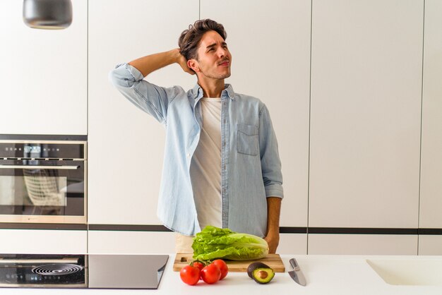 Jonge man van gemengd ras die een salade bereidt voor de lunch, achterhoofd aanraakt, nadenkt en een keuze maakt.