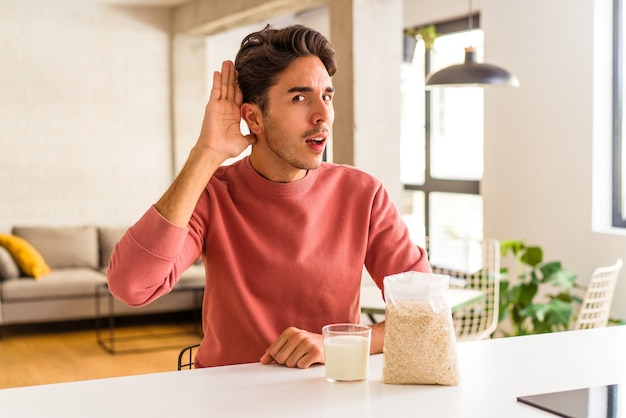 Jonge man van gemengd ras die havermout en melk eet als ontbijt in zijn keuken en probeert te luisteren naar roddels.