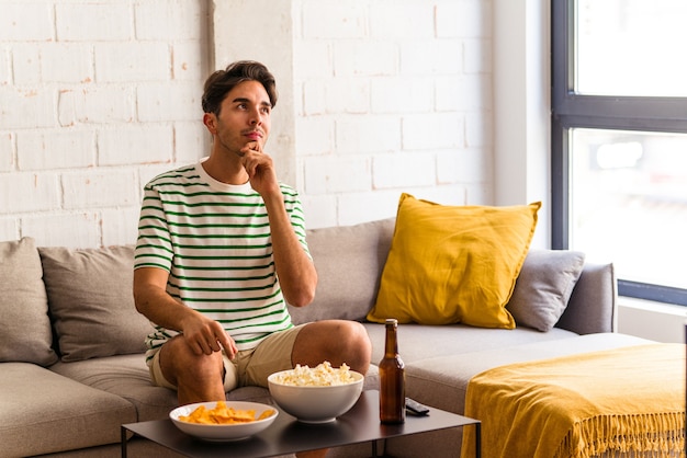 Jonge man van gemengd ras die popcorn eet terwijl hij op de bank zit en zijwaarts kijkt met een twijfelachtige en sceptische uitdrukking.