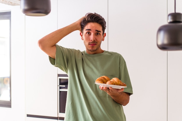 Jonge man van gemengd ras die 's ochtends een croissant in een keuken eet en geschokt is, herinnert zich een belangrijke ontmoeting.