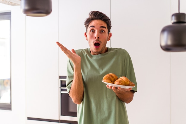 Jonge man van gemengd ras die 's ochtends een croissant in een keuken eet, verrast en geschokt.