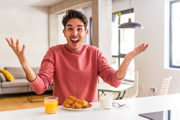 Jonge man van gemengd ras die 's ochtends ontbijt in een keuken en een aangename verrassing ontvangt, opgewonden en handen opsteekt.