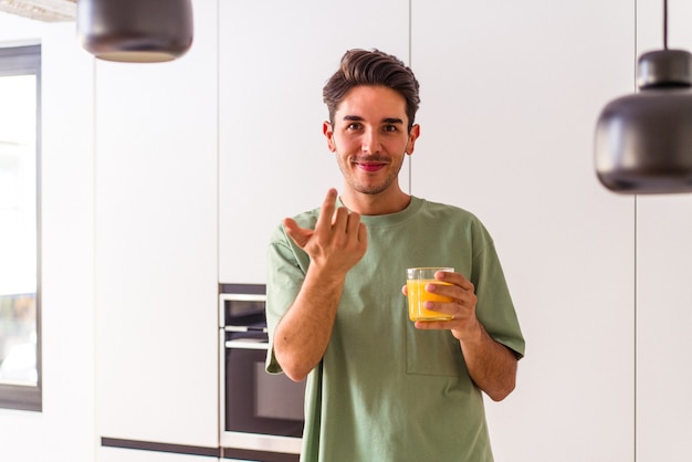 Jonge man van gemengd ras die sinaasappelsap drinkt in zijn keuken en met de vinger naar je wijst alsof hij dichterbij komt.