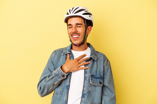 Jonge man van gemengd ras met een helmfiets geïsoleerd op gele achtergrond lacht hardop terwijl hij de hand op de borst houdt.