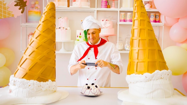 Jonge man verkleed als chef-kok die cake op mobiele telefoon in zoetwaren fotografeert Mannelijke banketbakker in uniform met groene dreadlocks die foto's maken van zijn koken op gadget