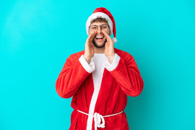 Jonge man vermomd als kerstman geïsoleerd op blauwe achtergrond schreeuwen en iets aankondigen
