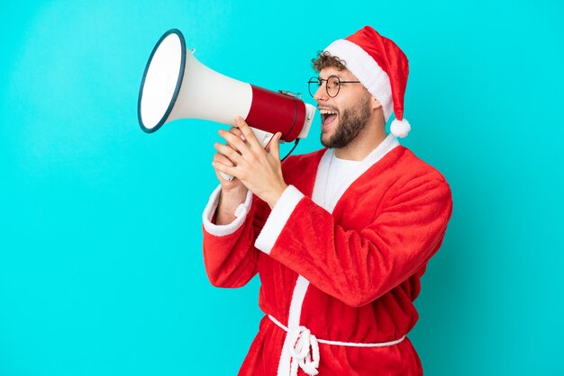 Jonge man vermomd als kerstman geïsoleerd op blauwe achtergrond schreeuwend door een megafoon