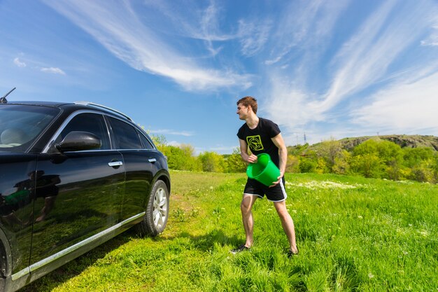 Jonge man wassen zwarte luxevoertuig in grasachtig groen veld op zonnige dag met blauwe lucht, emmer water aan de zijkant van de auto gooien