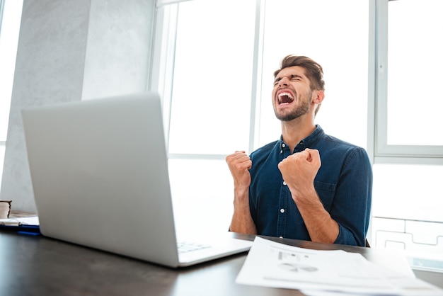 Jonge man winnaar gebaar maken zittend aan tafel en laptop