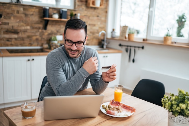 Jonge man zit aan de keukentafel met ontbijt in de ochtend met behulp van laptop en creditcard.