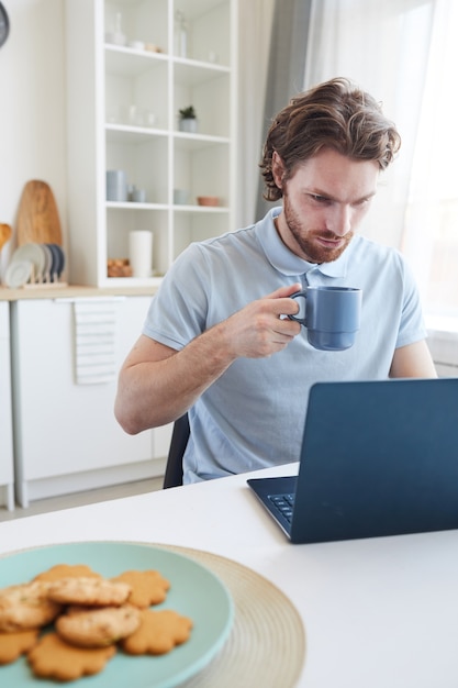 Jonge man zit aan de tafel die op laptop werkt en koffie drinkt in de ochtend in de keuken