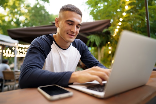 Jonge man zit aan tafel en typt op het toetsenbord van de laptop terwijl hij op het terras werkt