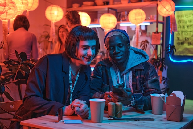 Jonge man zit aan tafel met zijn vriendje, ze kijken naar iets op een mobiele telefoon in een nachtclub