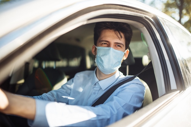 Jonge man zit achter het stuur in de auto met een steriel medisch masker. Sociale afstand, virusverspreiding preventie en behandelconcept.