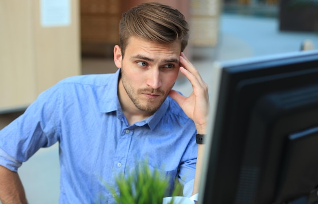 Foto jonge man zit en kijkt naar computermonitor terwijl hij op kantoor werkt.