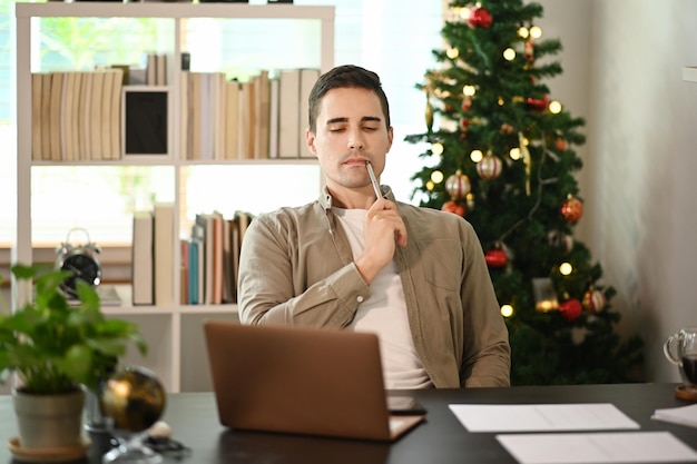 Jonge man zit in de buurt van de kerstboom en gebruikt een laptopcomputer