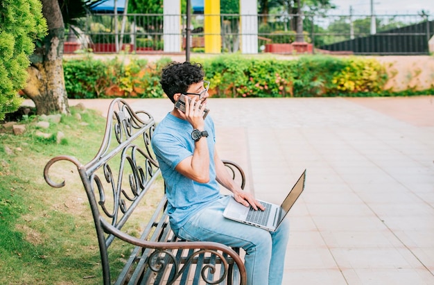 Jonge man zit in een park aan het werk met zijn laptop en mobiele telefoon Man in een park werkt online met laptop en belt op mobiele telefoon Freelancer man aan het werk met laptop terwijl hij telefoneert