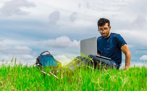 Jonge man zit met laptop en rugzak in het veld Man zit in het groene veld werken vanaf zijn laptop Freelancer man concept werken vanuit het veld