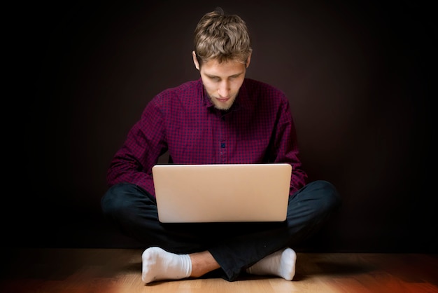 Jonge man zit op de vloer en gebruikt een laptop op een donkere achtergrond b