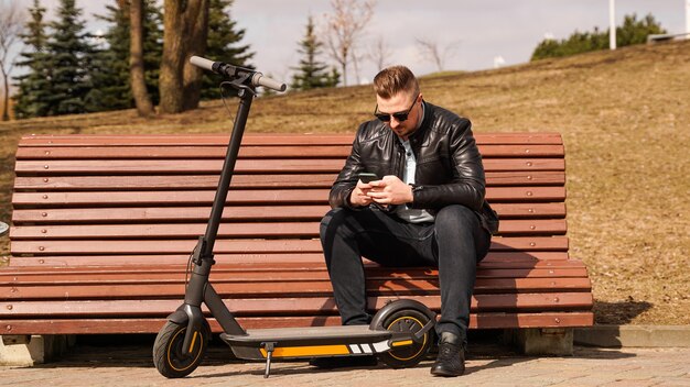 Jonge man zit op een bankje op een elektrische scooter van de lentedag naast een bankje