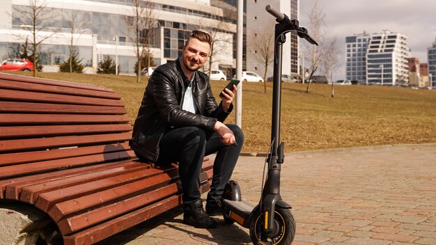 Jonge man zit op een bankje op een elektrische scooter van de lentedag naast een bankje