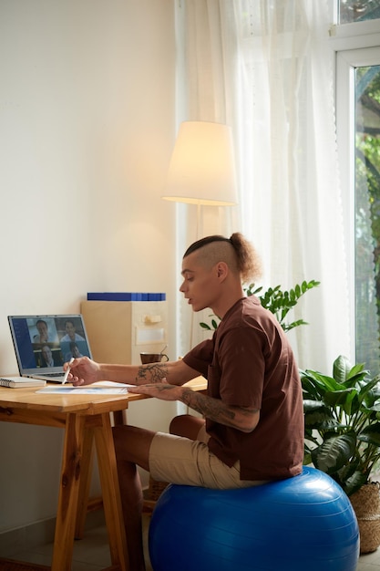 Jonge man zit op fitnessbal bij het bespreken van het rapport met collega's tijdens online vergadering