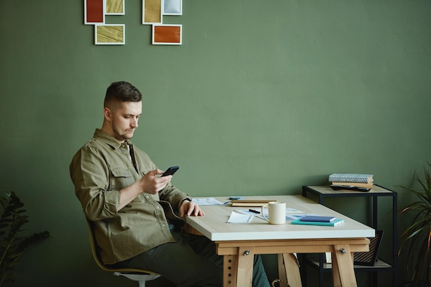 Jonge man zit op zijn werkplek thuis en typt een bericht op mobiele telefoon die hij online werk doet