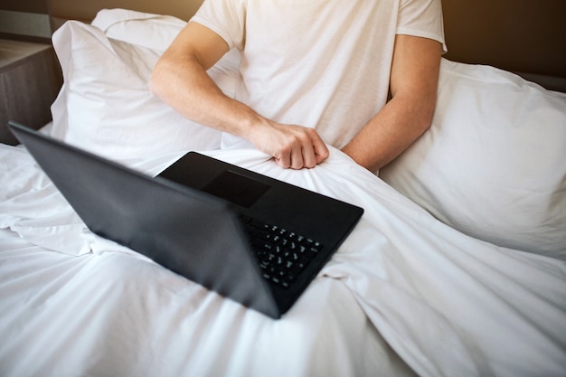 Jonge man zit vroeg in de ochtend in bed. Hij houdt de hand onder een witte deken en masturbeert. Laptop op zijn benen. Video aan het kijken