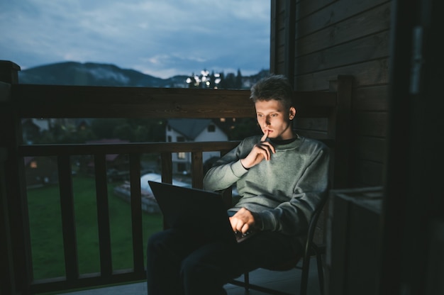 Jonge man zittend in een stoel op een balkon met behulp van een laptop